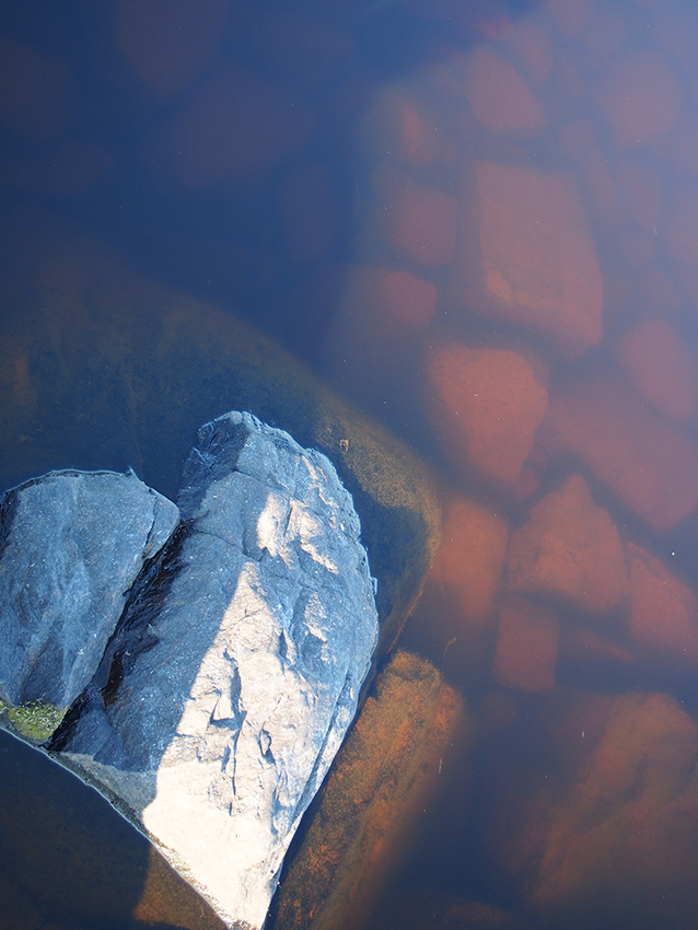 Veden pohjasta heijastuvia oranssin värisiä kiviä. Yksi iso kivi on veden pinnan yläpuolella ja muodosata sydän-kuvion.